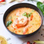 Thai coconut shrimp soup in a black bowl with creamy orange broth, topped with shrimp and herbs, with sliced lime, lemon, chili, and aromatics on a light surface.
