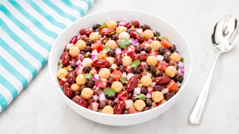 Editorial close-up of cowboy caviar in a white serving bowl with chickpeas, kidney beans, black beans, tomatoes, red onion, and cilantro, beside a teal-striped towel and silver spoon on a marble surface.