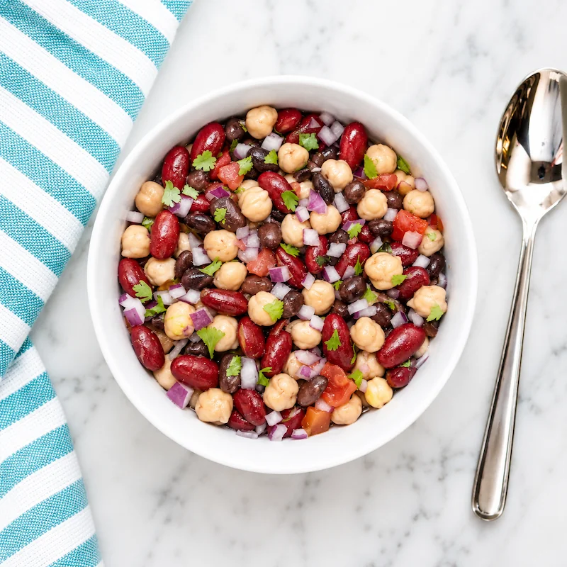 Aerial view of cowboy caviar in a white bowl with chickpeas, kidney beans, black beans, tomatoes, red onion, and cilantro, styled on white marble with a teal-and-white striped towel and silver spoon.
