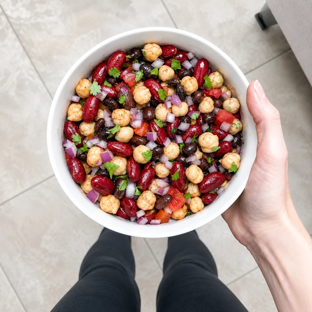 Cowboy Caviar Overhead view of a white bowl filled with cowboy caviar, featuring chickpeas, kidney beans, black beans, diced tomatoes, red onion, and cilantro, held above black leggings on a light tile floor.