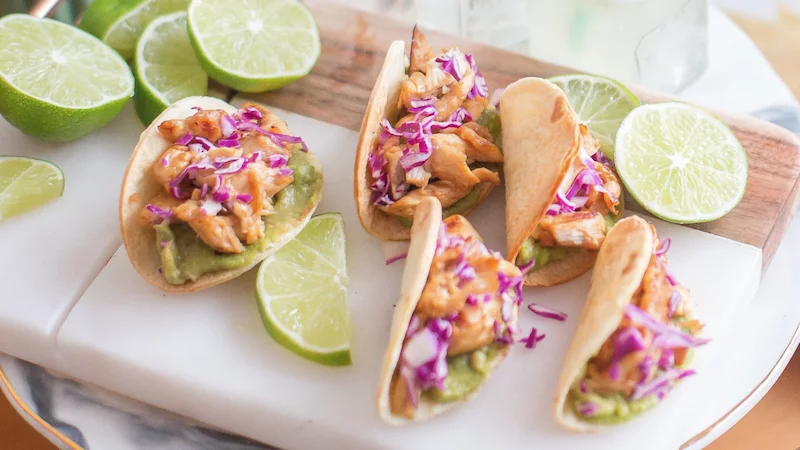 5 chicken, cabbage, and guacamole tacos on a cutting board with limes