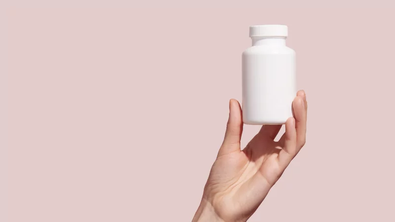 a hand holding a blank supplement bottle with a pink background