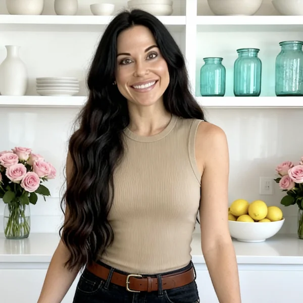 Dr. Christina Carlyle smiling in a bright kitchen, wearing a neutral sleeveless top and jeans, standing in front of shelves with glass jars, flowers, and fresh lemons, representing healthy lifestyle and functional nutrition expertise