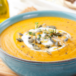 Close up of creamy butternut squash soup in a blue bowl topped with pumpkin seeds, herbs, and a swirl of cream on a wooden board.