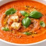 Close up of creamy tomato soup in a white bowl topped with basil leaves, croutons, olive oil, and black pepper on a wooden board.