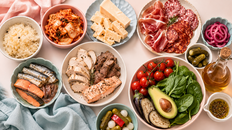 Flat lay of high histamine foods including fermented vegetables, kimchi, aged cheese, cured meats, canned fish, salmon, leftover meats, tomatoes, spinach, avocado, eggplant, pickles, vinegar, and vinegar-based dressing.