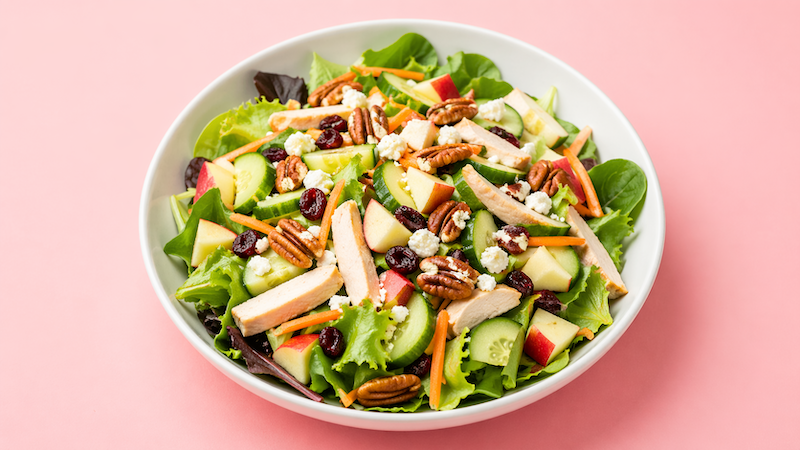 Apple pecan turkey summer salad with greens, cucumber, carrots, cranberries, and feta in a white bowl on a soft pink background