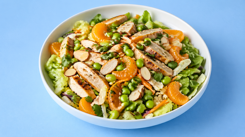 Asian chicken summer salad with mandarin oranges, edamame, shredded carrots, and sliced almonds in a white bowl on a light blue background