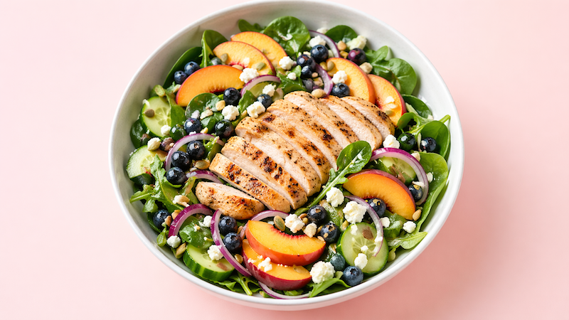 Peach blueberry chicken summer salad with mixed greens, cucumber, red onion, feta, and seeds in a white bowl on a light pink background