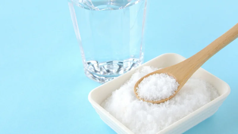 Close-up of a glass of water and a small dish of coarse white salt with a wooden spoon on a light blue background.