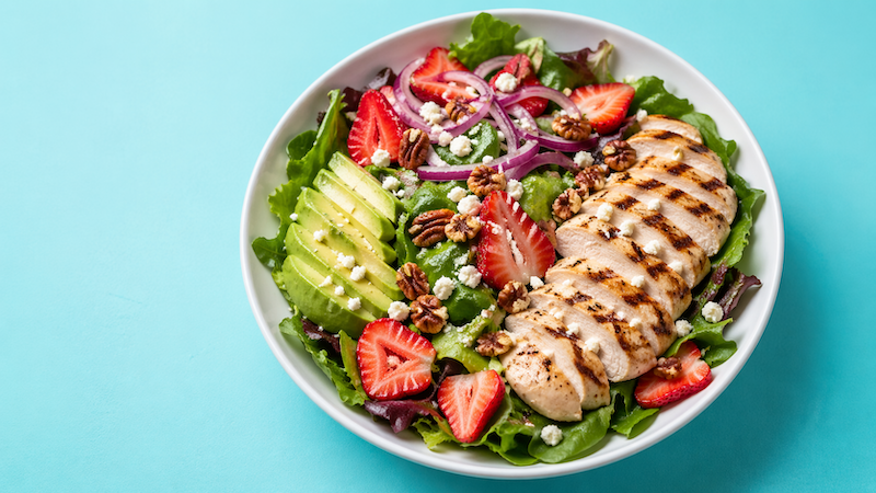 Strawberry avocado chicken summer salad with mixed greens, red onion, pecans, and feta in a white bowl on a teal background