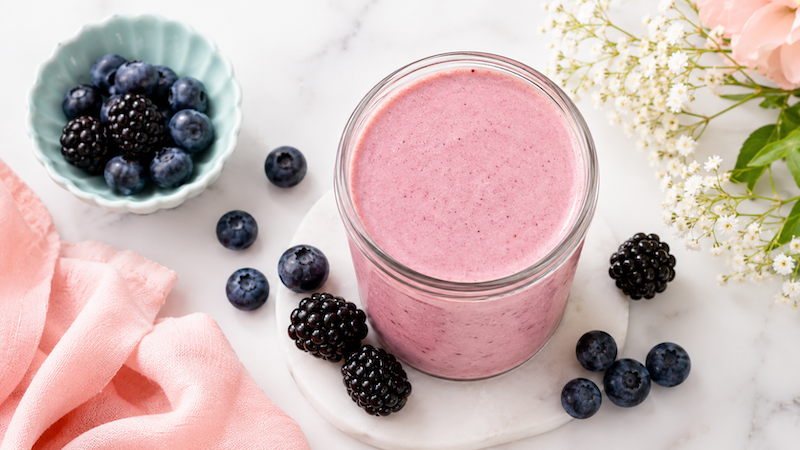 Berry Protein Smoothie Light berry protein smoothie in a glass jar with blueberries, blackberries, and soft pink wellness styling on a white marble background.