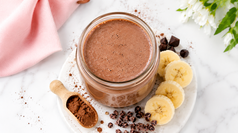 Chocolate Banana Metabolism Boosting Smoothie Chocolate banana breakfast smoothie in a glass jar with banana slices, cacao powder, chocolate pieces, and blush pink styling.