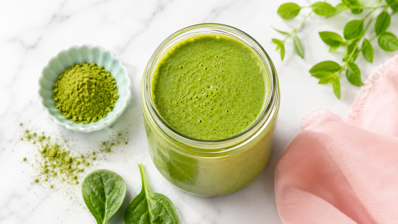 Green Power Shake Green superfood breakfast smoothie in a glass jar with green powder, leafy greens, and soft blush pink styling on a white marble background.