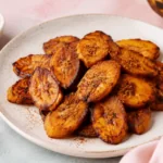 Fried sweet plantains on a white plate with cinnamon, a ripe plantain, pink napkin, and soft pastel food styling.