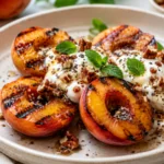 Grilled peaches topped with creamy yogurt, chopped pecans, chia seeds, honey, and fresh mint on a white plate for an easy healthy summer dessert.