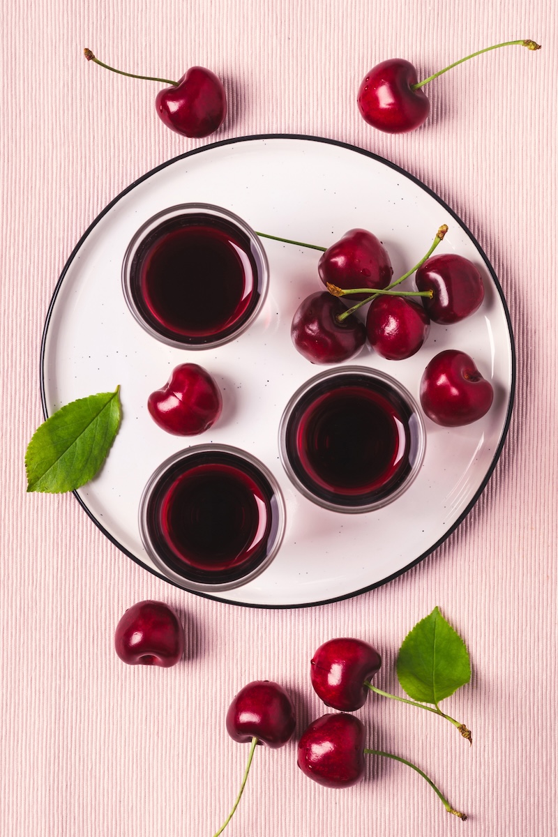 a flatlay of 3 glasses of tart cherry juice and organic cherries on a pink background